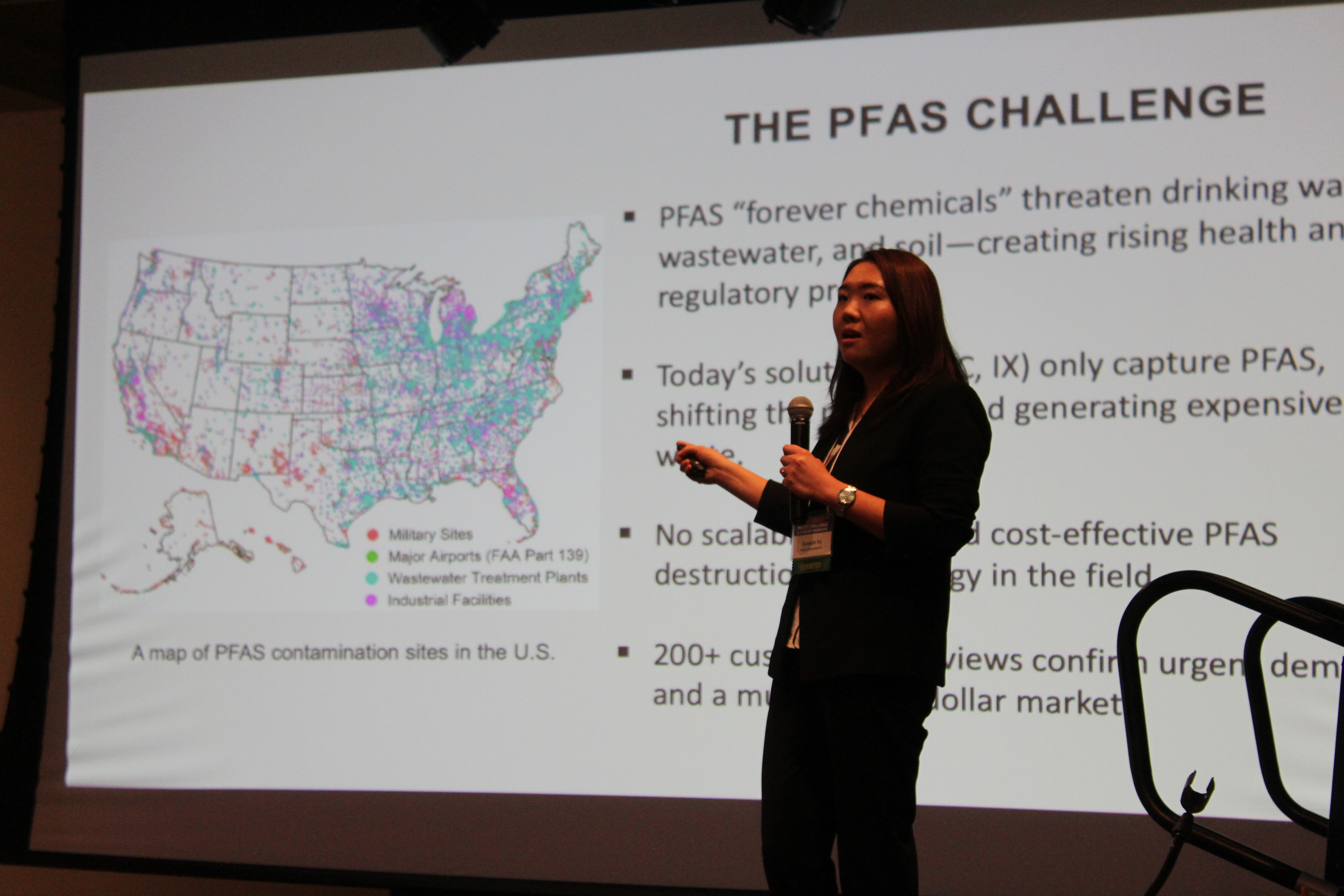 A woman on stage presenting her business pitch for her startup company in front of a large screen.